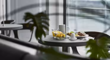 Et rundt bord med frokostartikler, inkludert en skål med frukt, et glass vann, en kaffekopp, en smørskål og et bakverk, sett gjennom grønne planteblader i en moderne spisestue med store vinduer.