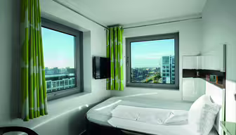 Et lite, moderne hotellrom med lyse grønne gardiner og to store vinduer med utsikt over byens bygninger. Rommet har en pent oppredd seng, en veggmontert TV og en elegant, minimalistisk design.