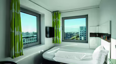 Et lite, moderne hotellrom med lyse grønne gardiner og to store vinduer med utsikt over byens bygninger. Rommet har en pent oppredd seng, en veggmontert TV og en elegant, minimalistisk design.