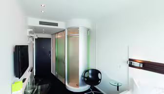 Moderne hotellrom med elegant design. Har en sirkulær glassdusj, veggmontert TV, svart svingstol, glassbord og en hvit seng. Rommet har svart teppe og hvite vegger med overlys.