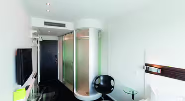 Moderne hotellrom med elegant design. Har en sirkulær glassdusj, veggmontert TV, svart svingstol, glassbord og en hvit seng. Rommet har svart teppe og hvite vegger med overlys.