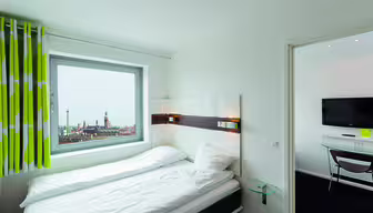 Et lyst hotellrom med en pent oppredd seng nær et vindu med utsikt over byen. Rommet har moderne design med veggmontert TV, grønne og hvite gardiner og et lite rundt glassbord ved siden av sengen.