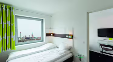 Et lyst hotellrom med en pent oppredd seng nær et vindu med utsikt over byen. Rommet har moderne design med veggmontert TV, grønne og hvite gardiner og et lite rundt glassbord ved siden av sengen.