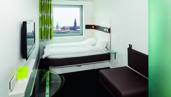 Lite hotellrom med en hvit seng ved siden av et vindu som viser et bybilde med tårn eller spir. Rommet har en brun sofa, en vegghengt TV og et glassbord med et gult hefte. Gardiner har et grønt og hvitt mønster.