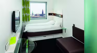 Lite hotellrom med en hvit seng ved siden av et vindu som viser et bybilde med tårn eller spir. Rommet har en brun sofa, en vegghengt TV og et glassbord med et gult hefte. Gardiner har et grønt og hvitt mønster.