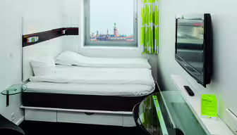 Et lite, moderne hotellrom med dobbeltseng ved siden av et vindu som viser et bybilde med et tårn. Rommet har vegghengt TV, skrivebord i glass og grønn gardin. Sengetøyet er hvitt, og gulvet er teppebelagt i mørke farger.