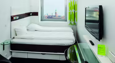 Et lite, moderne hotellrom med dobbeltseng ved siden av et vindu som viser et bybilde med et tårn. Rommet har vegghengt TV, skrivebord i glass og grønn gardin. Sengetøyet er hvitt, og gulvet er teppebelagt i mørke farger.