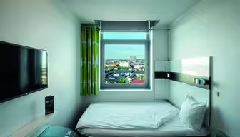 Et lite, moderne hotellrom med en enkeltseng ved et stort vindu som viser et bybilde. Rommet har vegghengt TV, grønnmønstret gardin og sidebord i glass. Vegger og sengetøy er hvite, og skaper et minimalistisk utseende.