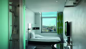 Moderne, kompakt hotellrom med enkeltseng, veggmontert TV og dusjkabinett i glass. Et vindu vender ut mot et bybilde med bygninger og kraner. Rommet har minimalistisk innredning og elegante møbler i grønn og hvit fargeskala.