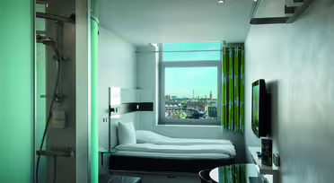 Moderne, kompakt hotellrom med enkeltseng, veggmontert TV og dusjkabinett i glass. Et vindu vender ut mot et bybilde med bygninger og kraner. Rommet har minimalistisk innredning og elegante møbler i grønn og hvit fargeskala.