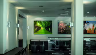 Et moderne kaféinteriør med svarte sitteplasser og små runde bord. Veggene er utsmykket med store bilder, inkludert et levende bilde av en trekantet sti. Kraftig belysning fremhever den rene og minimalistiske innredningen.