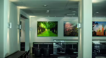 Et moderne kaféinteriør med svarte sitteplasser og små runde bord. Veggene er utsmykket med store bilder, inkludert et levende bilde av en trekantet sti. Kraftig belysning fremhever den rene og minimalistiske innredningen.
