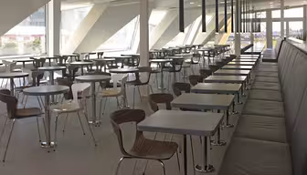 Romslig kafeteria med moderne design, med rader med firkantede bord med trestoler. Store vinduer gir naturlig lys til å fylle plassen, og en lang svart polstret benk linjer den ene siden.