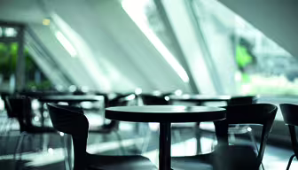 En moderne, tom kafé med elegante, mørke runde bord og stoler. Store vinduer lar naturlig lys strømme inn, og kaster geometriske skygger på gulvet. Atmosfæren er minimalistisk og rolig.
