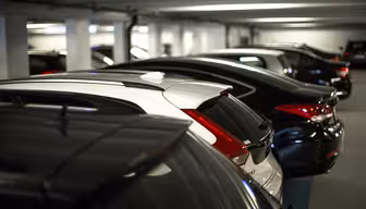En rad med parkerte biler i et svakt opplyst innendørs parkeringshus. Kjøretøyene er parallelt justert, med stort sett mørke farger, og én hvit bil skiller seg ut blant dem. Garasjen har betongsøyler og lavt tak med lysrør.