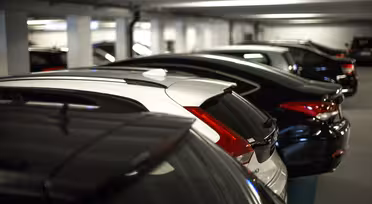 En rad med parkerte biler i et svakt opplyst innendørs parkeringshus. Kjøretøyene er parallelt justert, med stort sett mørke farger, og én hvit bil skiller seg ut blant dem. Garasjen har betongsøyler og lavt tak med lysrør.