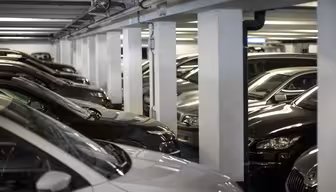 Et svakt opplyst underjordisk parkeringshus med flere biler parkert i parallelle rader. Hvite betongsøyler skiller kjøretøyene og skaper smale ganger. Rommet fremstår som rent og organisert, med en moderne industriell estetikk.