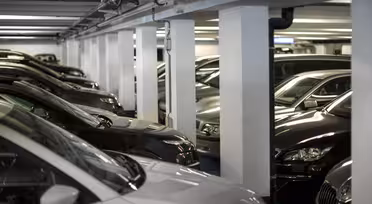 Et svakt opplyst underjordisk parkeringshus med flere biler parkert i parallelle rader. Hvite betongsøyler skiller kjøretøyene og skaper smale ganger. Rommet fremstår som rent og organisert, med en moderne industriell estetikk.