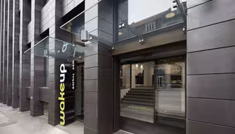 Utvendig visning av en moderne bygning med mørkt panel, med en baldakin i glass over en inngang. Inngangspartiet har store skyvedører i glass med teksten «wakeup copenhagen» på. Innvendig er det trappetrinn som fører opp til hovedområdet.