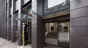 Utvendig visning av en moderne bygning med mørkt panel, med en baldakin i glass over en inngang. Inngangspartiet har store skyvedører i glass med teksten «wakeup copenhagen» på. Innvendig er det trappetrinn som fører opp til hovedområdet.