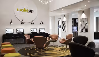 Moderne hotellobby med elegante oransje og svarte stoler rundt sirkulære mønstrede tepper. En rad med datastasjoner langs veggen under et stort skilt med teksten "wakeup aarhus.dk." Dekorativ belysning og speil forbedrer plassen.