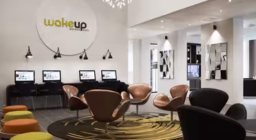 Moderne hotellobby med elegante oransje og svarte stoler rundt sirkulære mønstrede tepper. En rad med datastasjoner langs veggen under et stort skilt med teksten "wakeup aarhus.dk." Dekorativ belysning og speil forbedrer plassen.