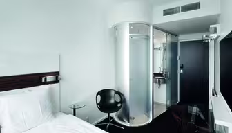 Et moderne hotellrom med en enkeltseng, en svart stol og et glassbord. Det er en unik sylindrisk dusj og et bad med glassvegger. Rommet er minimalistisk med hvite vegger og mørke aksenter. En flatskjerm-TV er montert på veggen.