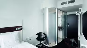 Et moderne hotellrom med en enkeltseng, en svart stol og et glassbord. Det er en unik sylindrisk dusj og et bad med glassvegger. Rommet er minimalistisk med hvite vegger og mørke aksenter. En flatskjerm-TV er montert på veggen.