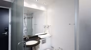 Moderne bad med dusjdør i glass, veggspeil, svart benkevask og hvitt toalett. Håndklær er pent brettet på benken. Designet er elegant med hvite vegger og en minimalistisk stil.