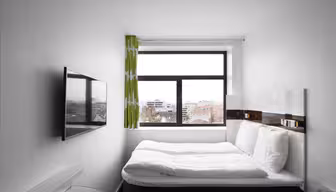 Et lite, minimalistisk soverom med en pent oppredd seng ved siden av et stort vindu med utsikt over bybildet. Veggene er hvite, og det er en flatskjerm-TV montert på veggen. Grønne gardiner henger ved siden av vinduet.