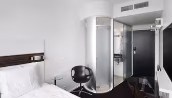Moderne hotellrom med hvit seng, svart stol og glassbord. Rommet har en unik baderomsdusj og dør i sylindrisk glass. Veggene er hvite, og det er en stor flatskjerm-TV montert på veggen.