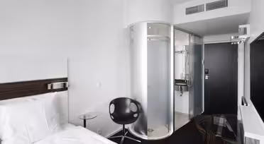 Moderne hotellrom med hvit seng, svart stol og glassbord. Rommet har en unik baderomsdusj og dør i sylindrisk glass. Veggene er hvite, og det er en stor flatskjerm-TV montert på veggen.