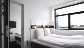 Et minimalistisk hotellrom med en stor seng utsmykket med hvitt sengetøy, en veggmontert TV og et vindu med utsikt over byen. En annen seng er synlig gjennom en åpen dør til et sammenhengende rom.