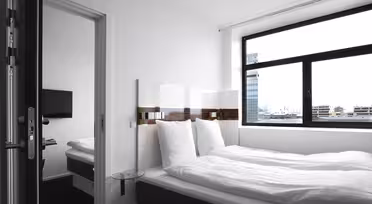 Et minimalistisk hotellrom med en stor seng utsmykket med hvitt sengetøy, en veggmontert TV og et vindu med utsikt over byen. En annen seng er synlig gjennom en åpen dør til et sammenhengende rom.