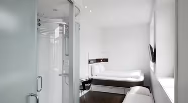 Minimalistisk hotellrom med seng, lite skrivebord med stol og veggmontert TV. En dør i frostet glass fører til et kompakt dusjområde. Rommet er lyst med naturlig lys fra et stort vindu, med hvite vegger og mørkt tregulv.