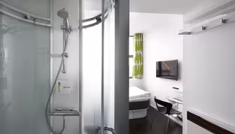 Moderne hotellrom med en buet glassdusj til venstre og en liten stue til høyre. Rommet har en veggmontert TV, en seng med hvitt sengetøy, et skrivebord med glassoverflate, en svart stol og et vindu med grønne mønstrede gardiner.