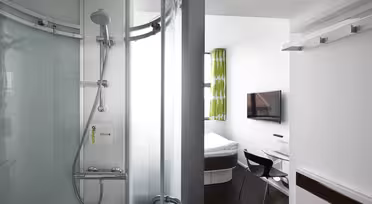 Moderne hotellrom med en buet glassdusj til venstre og en liten stue til høyre. Rommet har en veggmontert TV, en seng med hvitt sengetøy, et skrivebord med glassoverflate, en svart stol og et vindu med grønne mønstrede gardiner.