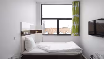 Lite, moderne soverom med hvite vegger med en enkeltseng med hvitt sengetøy. Det er et stort vindu med grønne gardiner mønstret med hvitt. En flatskjerm-TV er montert på veggen, med et sidebord med glassplate i nærheten.
