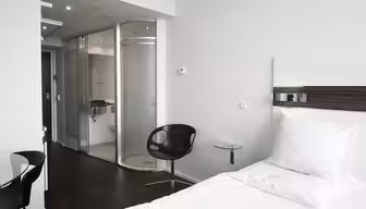 Moderne hotellrom med minimalistisk design. Har en hvit enkeltseng, svart svingstol og et lite sidebord i glass. Badet har glassdør og elegant inventar. Rommet har en nøytral fargepalett med hvite vegger og mørkt gulv.