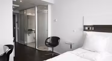 Moderne hotellrom med minimalistisk design. Har en hvit enkeltseng, svart svingstol og et lite sidebord i glass. Badet har glassdør og elegant inventar. Rommet har en nøytral fargepalett med hvite vegger og mørkt gulv.