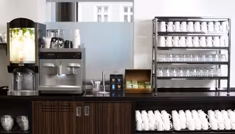 En selvbetjent kaffestasjon med ulike kaffemaskiner på disk. I nærheten holder stativer pent stablede hvite kopper og glass. En boks med teposer er synlig, og omgivelsene er lyse med naturlig lys fra vinduene.