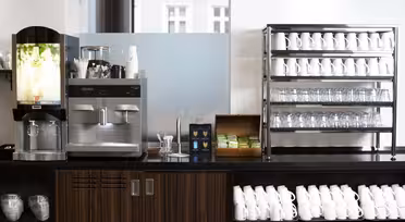 En selvbetjent kaffestasjon med ulike kaffemaskiner på disk. I nærheten holder stativer pent stablede hvite kopper og glass. En boks med teposer er synlig, og omgivelsene er lyse med naturlig lys fra vinduene.