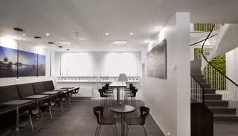 Moderne kafé med minimalistisk design, med svarte stoler og bord på mørke flislagte gulv. Store veggutskrifter og en trapp med grøntområder legger til dekor. Hvite vegger og taklys skaper en lys, åpen atmosfære.