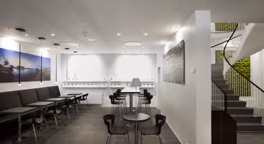 Moderne kafé med minimalistisk design, med svarte stoler og bord på mørke flislagte gulv. Store veggutskrifter og en trapp med grøntområder legger til dekor. Hvite vegger og taklys skaper en lys, åpen atmosfære.