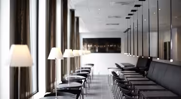 Et moderne, minimalistisk kaféinteriør med rader med små, runde sorte bord og stoler foret med hvite lamper. Store vinduer til venstre og speilvegg til høyre forsterker rommets lysstyrke.