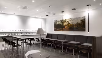 Et moderne, minimalistisk kaféinteriør med grå vegger og gulv. Langbord omkranset av trestoler og høye krakker. Et stort plantebilde dekorerer veggen over en polstret benk. Lyse taklamper forsterker den rene, åpne plassen.