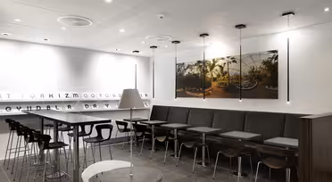 Et moderne, minimalistisk kaféinteriør med grå vegger og gulv. Langbord omkranset av trestoler og høye krakker. Et stort plantebilde dekorerer veggen over en polstret benk. Lyse taklamper forsterker den rene, åpne plassen.