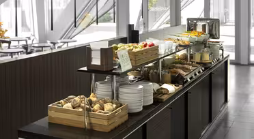 En frokostbuffé med rundstykker, skåler med frukt, stablede tallerkener, ulike pålegg, en brødrister og andre frokostartikler. Innstillingen er moderne med naturlig lys som kommer inn gjennom store vinduer.
