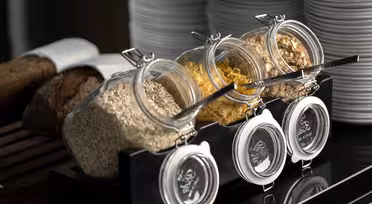 Tre glasskrukker er fylt med havre, cornflakes og granola, hver med en skje. De vises på et svart stativ. Stabler med hvite tallerkener og brød er i bakgrunnen. Innstillingen foreslår en frokostbuffé.