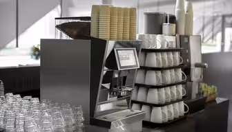 Et moderne kaffemaskinoppsett med stabler av papirkopper på toppen. Glass og hvite keramiske kopper er pent arrangert i hyller. En organisert visning av tepakker er sett i nærheten, mot et bakteppe av et lyst, luftig rom.